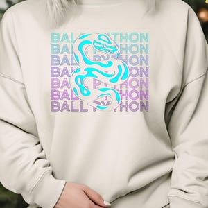 K&ouml;nnte beinhalten: Cremefarbenes Sweatshirt mit einem t&uuml;rkisfarbenen und wei&szlig;en Schlangenmuster und dem Text "BALL PYTHON" in einem sich wiederholenden Muster. Die Schlange ist stilisiert und der Text hat einen blauen und violetten Farbverlauf.
