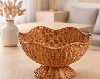 May include: A woven wicker basket with a scalloped rim, set on a pedestal base. The basket is a warm brown color and appears to be empty. The basket is sitting on a light-colored wooden surface.