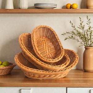 May include: Four woven wicker baskets of varying sizes are stacked on a wooden countertop. The baskets are oval-shaped with a light brown color. A small basket of green fruit is in the background.