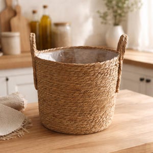 May include: A round, woven basket with two handles. The basket is made of natural-colored fibers and has a clear plastic liner. It sits on a light-colored wooden surface, suggesting a decorative or storage item.