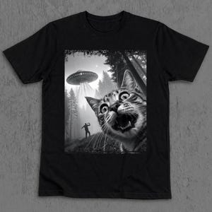 Könnte beinhalten: Schwarzes T-Shirt mit einer Schwarz-Weiß-Grafik, die eine überraschte Katze mit weit aufgerissenen Augen und offenem Mund zeigt. Ein außerirdisches Raumschiff strahlt Licht auf eine Person in einem Wald.