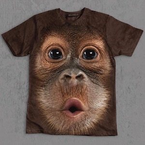 Può includere: Una t-shirt marrone a maniche corte con una grande stampa grafica realistica e ad alta definizione del volto di un orangotango. Il primate presenta grandi occhi scuri, un naso strutturato e labbra increspate su tutta la parte anteriore.