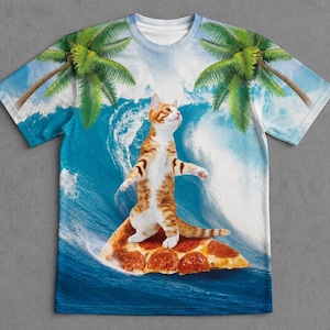 Op de afbeelding: Een t-shirt met korte mouwen en een all-over print van een oranje cyperse kat die op een punt pepperoni-pizza staat. De achtergrond toont een grote brekende oceaangolf met groene palmbomen die vanuit de bovenhoeken van het shirt uitsteken.