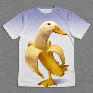 May include: A short-sleeve t-shirt featuring a graphic print of a banana shaped like a duck. The shirt has a light purple to white gradient background. The banana duck has a yellow peel, a yellow beak, and yellow webbed feet standing upright.
