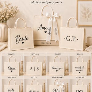 May include: Three off-white canvas tote bags with top handles. The center bag features a white satin ribbon and the text 'Ame', 'Amelia', and '09.12.25'. The left bag says 'Bride' with a heart, and the right bag says 'G.T.' with dots.