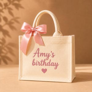 May include: A small beige canvas tote bag featuring two top handles and a large pink satin bow attached to the left side. The front of the bag displays the text Amy's birthday in pink glittery script lettering, followed by a small pink glittery heart.