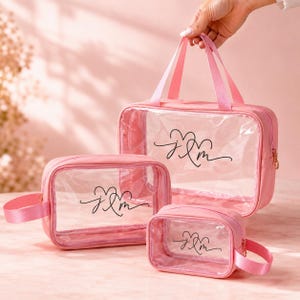 May include: Three pink cosmetic bags of varying sizes. Each bag has a clear front panel with a black script design. The largest bag has handles, while the other two have a handle or loop. The bags are zippered and appear to be made of a soft, flexible material.
