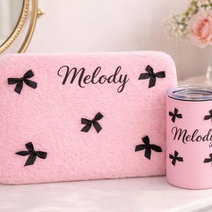May include: A pink, fluffy rectangular pillow and a matching tumbler, both adorned with black bow accents. The name "Melody" is scripted on both items. The tumbler has a silver lid. The items are on a white surface.