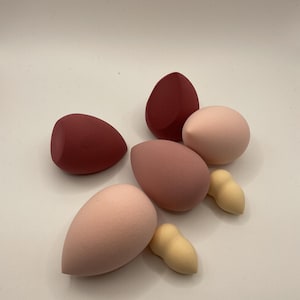 Puede incluir: Una colección de esponjas de maquillaje en varias formas y colores. Las esponjas incluyen formas de lágrima, huevo y cacahuete en tonos burdeos, rosa y amarillo. Los artículos están dispuestos sobre una superficie blanca.