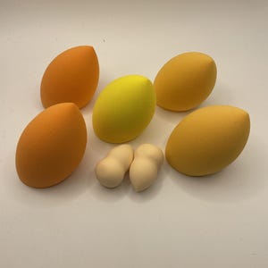 Puede incluir: Una selección de esponjas de maquillaje de varias formas y colores. La colección incluye seis esponjas en forma de huevo en tonos naranja, amarillo y beige claro. Estos aplicadores cosméticos están diseñados para difuminar el maquillaje.