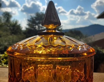 May include: A vintage amber glass candy dish with a diamond pattern and a pointed lid. The dish is sitting on a wooden surface.