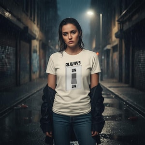 Pode incluir: Uma camiseta creme com as palavras "ALWAYS ON" acima de um gráfico de um interruptor na posição "ON". A camiseta é usada com jeans azuis e uma jaqueta preta. O fundo é uma cena de rua chuvosa.
