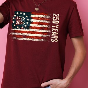 Puede incluir: Camiseta burdeos con un dise&ntilde;o desgastado de la bandera estadounidense. La bandera incluye los a&ntilde;os 1776 y 2026, con el texto "250 YEARS" impreso verticalmente. La camiseta es de manga corta y cuello redondo.