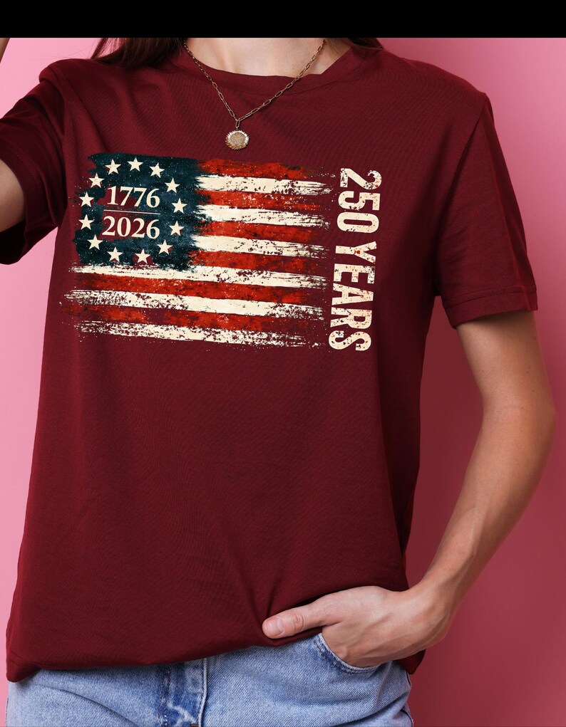 Puede incluir: Camiseta burdeos con un dise&ntilde;o desgastado de la bandera estadounidense. La bandera incluye los a&ntilde;os 1776 y 2026, con el texto "250 YEARS" impreso verticalmente. La camiseta es de manga corta y cuello redondo.