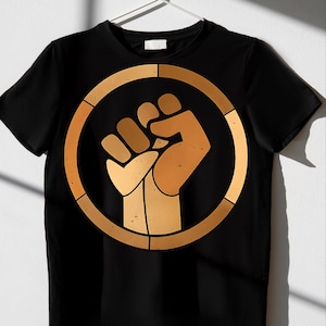 May include: Black t-shirt featuring a gold-colored fist inside a gold circle. The fist is raised in a symbol of solidarity. The design is centered on the front of the shirt, which is hanging on a white hanger.