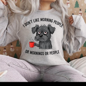 Könnte beinhalten: Ein hellgraues Sweatshirt mit dem Text "I DON'T LIKE MORNING PEOPLE OR MORNINGS OR PEOPLE." Die Grafik zeigt einen grummeligen schwarzen Hund, der eine rote Tasse hält. Das Sweatshirt ist ein lässiger, bequemer Stil.