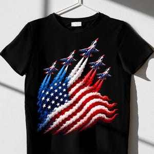 Könnte beinhalten: Ein schwarzes T-Shirt mit kurzen Ärmeln und einer Grafik von fünf Kampfjets in Formation. Die Jets hinterlassen Rauchfahnen in Blau, Weiß und Rot, die das Muster der amerikanischen Flagge bilden, einschließlich weißer Sterne auf blauem Grund.