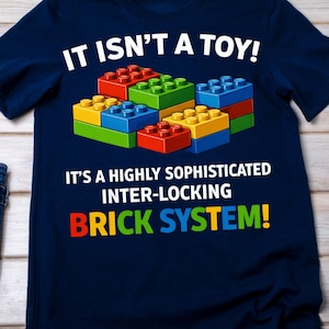 May include: Navy blue t-shirt with the text "IT ISN'T A TOY!" and "IT'S A HIGHLY SOPHISTICATED INTER-LOCKING BRICK SYSTEM!". The shirt features a graphic of colorful interlocking toy bricks in red, yellow, green, and blue.