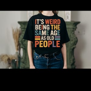 Puede incluir: Camiseta negra con el texto "IT'S WEIRD BEING THE SAME AGE AS OLD PEOPLE" en un esquema de colores retro. El texto está dispuesto en formato apilado. La persona lleva vaqueros azules.
