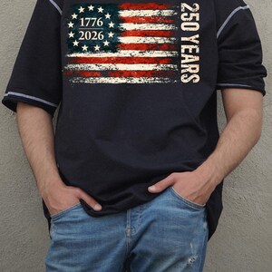 Puede incluir: Una camiseta azul marino de manga corta con un dise&ntilde;o de bandera americana desgastada, el texto "1776 2026" y "250 YEARS". La camiseta tiene costuras y ribetes blancos.