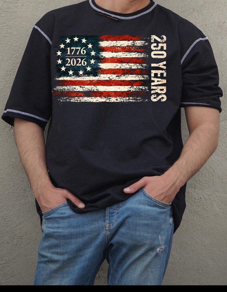 Puede incluir: Una camiseta azul marino de manga corta con un dise&ntilde;o de bandera americana desgastada, el texto "1776 2026" y "250 YEARS". La camiseta tiene costuras y ribetes blancos.