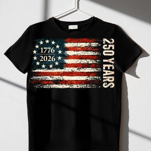 Puede incluir: Camiseta negra con un dise&ntilde;o desgastado de la bandera estadounidense. El dise&ntilde;o incluye el texto "1776 2026" y "250 YEARS". La bandera presenta rayas rojas y blancas, y un campo azul con estrellas blancas.