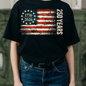 Puede incluir: Camiseta negra con un dise&ntilde;o desgastado de la bandera estadounidense. El dise&ntilde;o incluye el texto "1776 2026" y "250 YEARS" en blanco. El dise&ntilde;o de la bandera presenta rayas rojas y blancas y un c&iacute;rculo de estrellas.