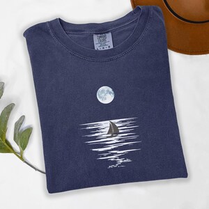 May include: A navy blue crew neck t-shirt featuring a graphic print of a full moon above a dark sailboat silhouette, with white horizontal lines representing light reflecting on water. The inner neck label displays the text Comfort Colors L-G-G.
