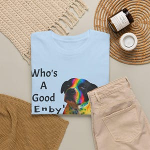 Peut inclure: T-shirt bleu clair avec le texte "Who's A Good Enby'" et un dessin de chien aux couleurs de l'arc-en-ciel. Le t-shirt est présenté avec un pantalon beige et un tapis tissé.