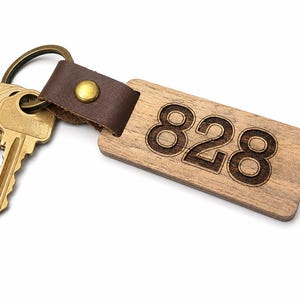 Könnte beinhalten: Ein Holzschlüsselanhänger mit der eingravierten Nummer "828" ist an einem braunen Lederriemen mit einem goldenen Niet befestigt. Der Schlüsselanhänger ist mit einem goldfarbenen Metallring verbunden, der drei goldene Schlüssel hält.