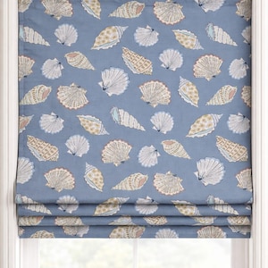 Puede incluir: Estor romano azul con un patrón repetido de conchas marinas blancas, beige y melocotón. El tratamiento de la ventana está montado dentro de un marco de ventana blanco, con un tema costero.