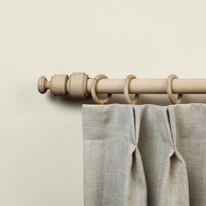 May include: A light-coloured wooden curtain rod with a decorative finial end piece, featuring three matching wooden rings suspended from the rod. A textured, light grey fabric curtain with vertical pleats hangs from the rings on the rod.