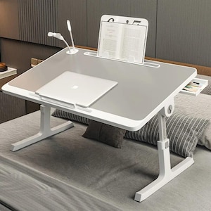 Op de afbeelding: Een witte en grijze verstelbare laptoptafel met een ingebouwde boekenstandaard en een kleine lade. Een zilveren laptop en een boek liggen op de tafel. De tafel heeft een kleine, verstelbare lamp. De tafel staat op een bed.
