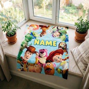 May include: A light blue rectangular fleece blanket featuring a colorful print of various Toy Story characters including Woody, Jessie, Buzz Lightyear, Rex, and Lotso. The word NAME is printed in large yellow block letters in the center.