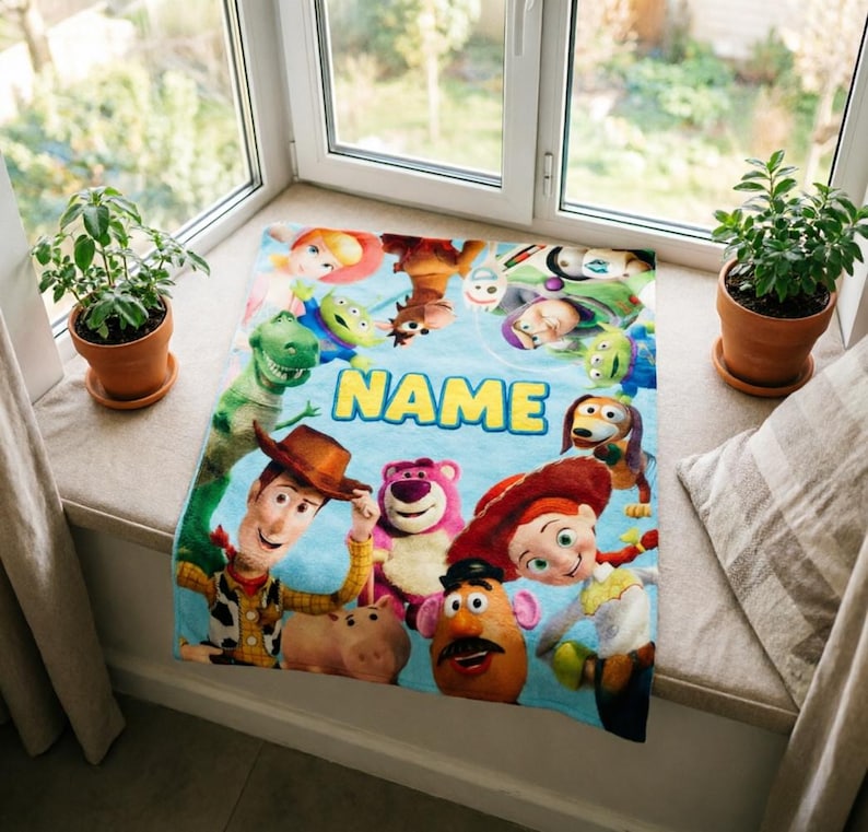 May include: A light blue rectangular fleece blanket featuring a colorful print of various Toy Story characters including Woody, Jessie, Buzz Lightyear, Rex, and Lotso. The word NAME is printed in large yellow block letters in the center.