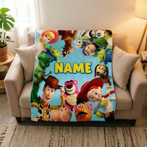 May include: A light blue fleece blanket featuring a colorful collage of Toy Story characters including Woody, Buzz Lightyear, Jessie, Rex, and Mr. Potato Head, with the word NAME printed in large yellow block letters in the center of the design.