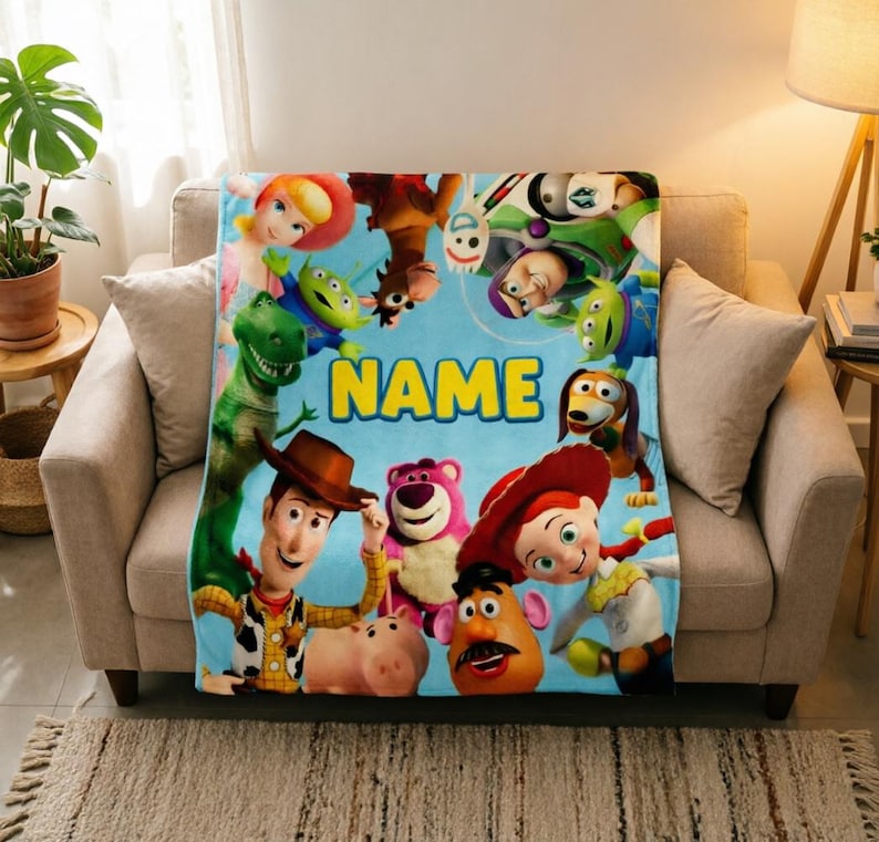 May include: A light blue fleece blanket featuring a colorful collage of Toy Story characters including Woody, Buzz Lightyear, Jessie, Rex, and Mr. Potato Head, with the word NAME printed in large yellow block letters in the center of the design.