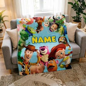May include: A light blue fleece blanket featuring a colorful collage of Toy Story characters including Woody, Jessie, Buzz Lightyear, Rex, and Forky, with the word NAME printed in large yellow block letters across the center of the design.