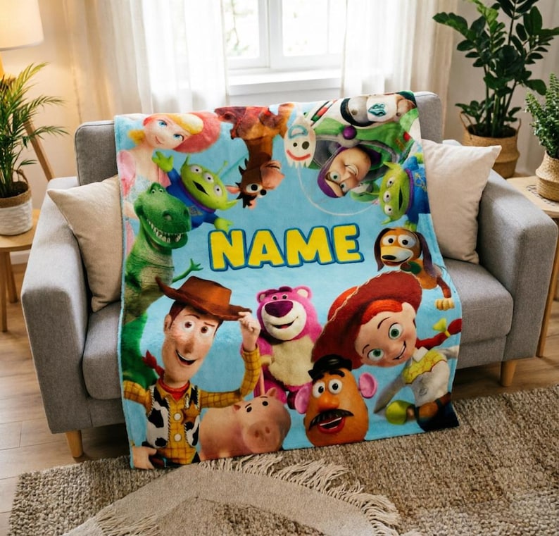 May include: A light blue fleece blanket featuring a colorful collage of Toy Story characters including Woody, Jessie, Buzz Lightyear, Rex, and Forky, with the word NAME printed in large yellow block letters across the center of the design.