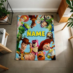 May include: A rectangular fleece blanket featuring a light blue background with a collage of various animated characters including a cowboy, a cowgirl, a green dinosaur, a pink bear, and a potato figure, with the word NAME printed in yellow.