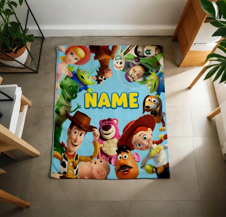 May include: A rectangular fleece blanket featuring a light blue background with a collage of various animated characters including a cowboy, a cowgirl, a green dinosaur, a pink bear, and a potato figure, with the word NAME printed in yellow.