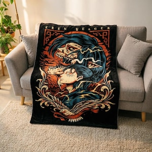 May include: A black rectangular throw blanket featuring a graphic illustration of Eren Yeager with a titan form above, orange and blue color accents, and a bird at the bottom. The text EREN YEAGER is printed in a stylized font at the top of the design.