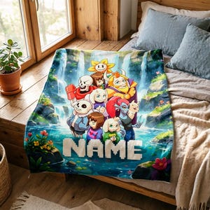 Op de afbeelding: Een blauwe deken met een cartoon illustratie van personages voor een waterval. De deken heeft het woord "NAME" in witte blokletters. De personages zijn in verschillende kleuren, waaronder rood, geel en blauw. De deken ligt op een bed.