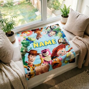 May include: A soft, light blue fleece blanket featuring a colorful collage of various Toy Story characters, including Woody, Jessie, Rex, and Lotso, arranged around the central yellow text that reads NAME in a bold, sans-serif font.