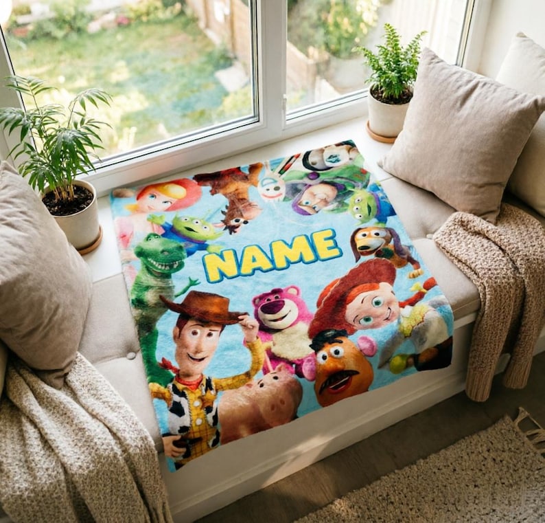 May include: A soft, light blue fleece blanket featuring a colorful collage of various Toy Story characters, including Woody, Jessie, Rex, and Lotso, arranged around the central yellow text that reads NAME in a bold, sans-serif font.