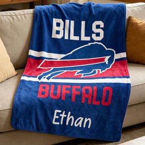 May include: A blue fleece blanket with the words "BILLS" and "BUFFALO" in white and red, along with a buffalo logo. The name "Ethan" is printed in white script. The blanket is draped over a beige sofa.