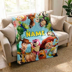 May include: A light blue fleece blanket featuring a colorful collage of Toy Story characters including Woody, Buzz Lightyear, Jessie, Rex, and Lotso. The word NAME is printed in large yellow block letters in the center of the blanket design.