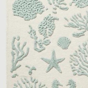 Puede incluir: Una alfombra rectangular con fondo color crema y un patr&oacute;n repetitivo de dise&ntilde;os en relieve de color azul claro. Los motivos incluyen varios tipos de corales, estrellas de mar, conchas marinas y caballitos de mar.