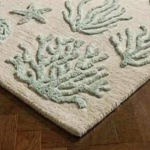 Puede incluir: Una alfombra rectangular con una base de color crema y un patr&oacute;n texturizado en relieve de vida marina en color verde espuma de mar claro, que incluye varias formaciones de coral y una estrella de mar, dispuestas en un dise&ntilde;o repetitivo.