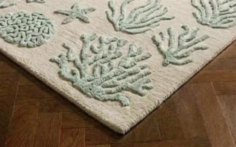 Puede incluir: Una alfombra rectangular con una base de color crema y un patr&oacute;n texturizado en relieve de vida marina en color verde espuma de mar claro, que incluye varias formaciones de coral y una estrella de mar, dispuestas en un dise&ntilde;o repetitivo.